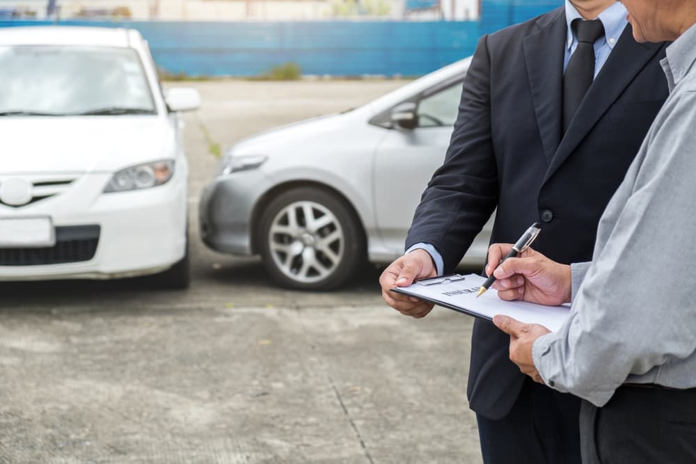 Insurance representative with victim standing next to a damaged car signing insurance claim documents in a parking lot.